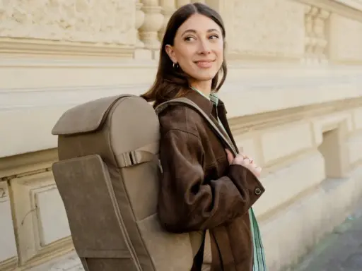 A woman smiles, showcasing the latest backpacks trend with a stylish, taupe backpack.
