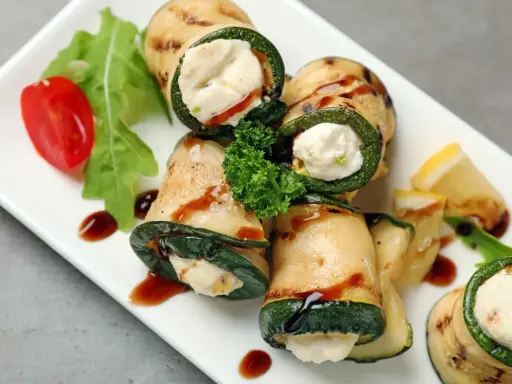 Deliziosi involtini di zucchine grigliate ripieni di formaggio cremoso, guarniti con pomodorini, rucola e salsa scura.