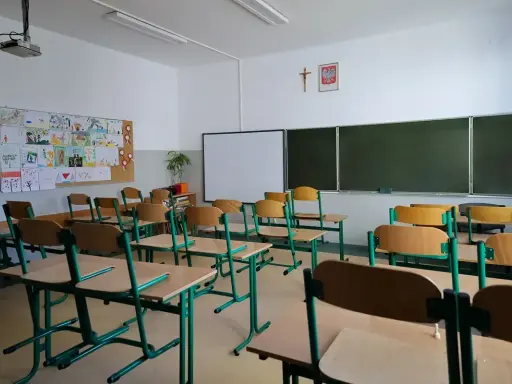 Pusta klasa z rzędami ławek i krzeseł, tablicą i dziecięcymi rysunkami na ścianie. Brak ucznia w tej sali może sugerować nieklasyfikowanie ucznia.