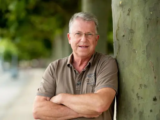 Carsten Sostmeier, ein Mann mit Brille und grauem Haar, lehnt lächelnd an einem Baum.