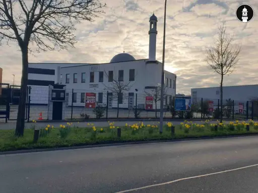 Moderne Subhan Moschee mit Minarett und Kuppel, umgeben von einem Zaun mit Schildern. Gelbe Narzissen blühen davor.