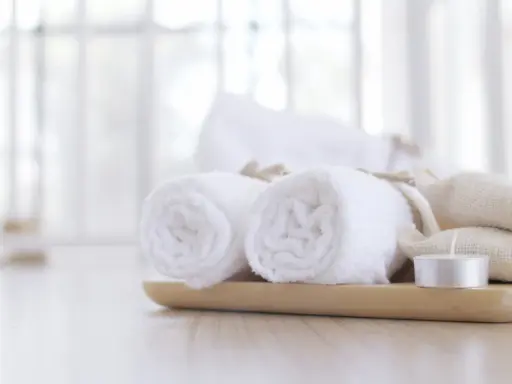 Lavette bianche arrotolate su un vassoio di legno, con sacchetti di tela e una candela. Un'immagine rilassante che evoca la cura di sé.