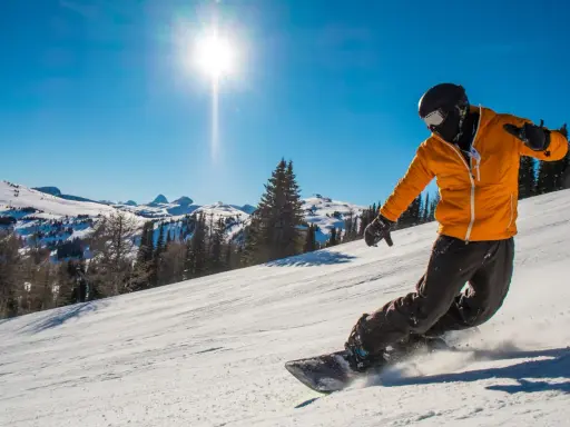 Snowboardzista w pomarańczowej kurtce zjeżdża po stoku, unosząc śnieg. To świetny przykład, jak jeździć na snowboardzie.