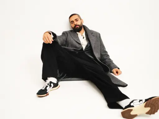 Ardalan Esmaili sitter avslappnat i en grå rock, vit skjorta och svarta byxor. Hans sneakers har en unik design.