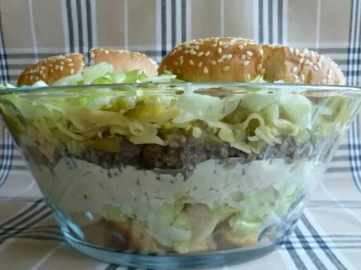 Warstwowa sałatka big mac w szklanej misie, z bułkami na wierzchu.