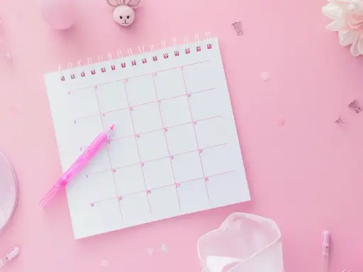 Różowy kalendarz z zaznaczonymi dniami, obok różowy pisak. Planowanie cyklu i test owulacyjny.