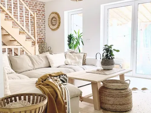 Jasny salon w stylu boho z beżową kanapą, drewnianymi schodami i ceglaną ścianą.