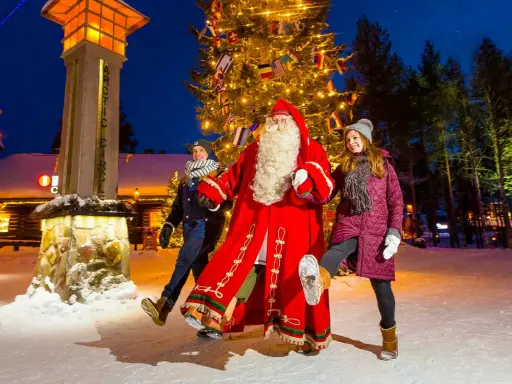 Santa Claus mit zwei Besuchern vor einem Weihnachtsbaum im Rovaniemi Weihnachtsdorf.
