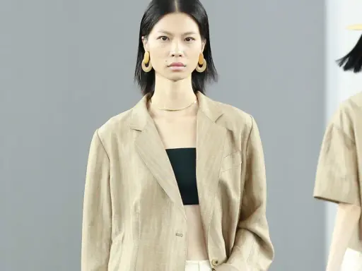 Ein Model trägt einen beigen Blazer aus knitterndem Leinen mit einem schwarzen Top. Große, runde Ohrringe schmücken ihre Ohren.