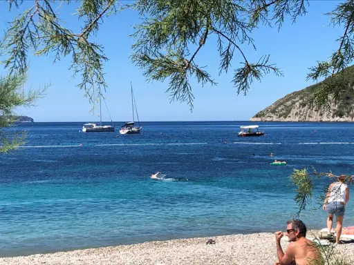 En man sitter på en stenstrand vid havet. I vattnet simmar en person och flera båtar ligger för ankar. En grön kulle reser sig i bakgrunden. En idyllisk vy från Vis, Kroatien.