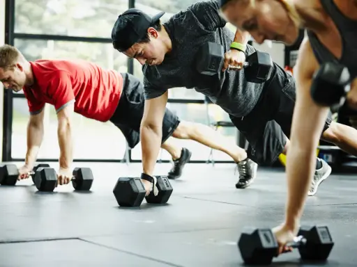 Drei Personen machen eine Plank-Übung mit Kurzhanteln. Ein Mann in Rot, ein Mann mit Kappe und eine Frau in Grau trainieren. CrossFit gesund und stark.