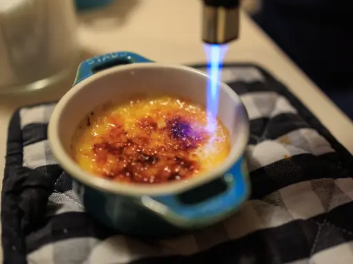 Flamme bleue caramélisant le sucre d'une crème brûlée, rappelant son origine.