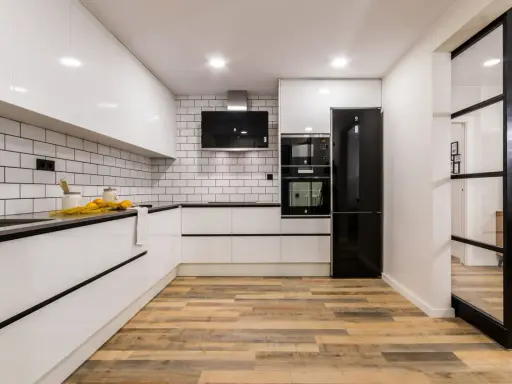 Cocina moderna con pared cocinas mitad azulejo mitad pintura blanca, electrodomésticos negros y suelo de madera.