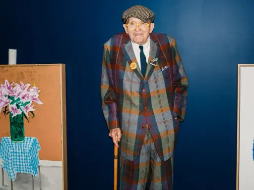 Ein Mann im karierten Anzug, inspiriert von David Hockney Pop Art, steht neben seinen lebendigen Gemälden.