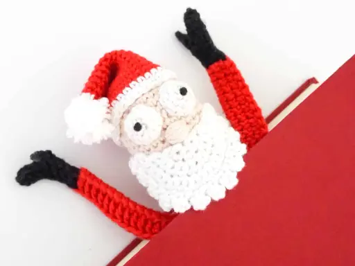 Marcador de libro amigurumi de Papá Noel, con brazos rojos y gorro festivo, asomando de un libro rojo.