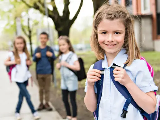 Uśmiechnięta dziewczynka z plecakiem, gotowa na zmianę szkoły średniej w 2 klasie technikum. W tle jej koledzy i koleżanki.