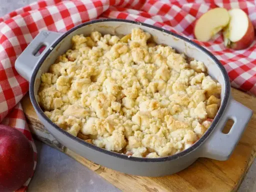 Ein goldbrauner Apfel Crumble in einer Auflaufform, bereit zum Genießen. Daneben liegen frische Äpfel auf kariertem Tuch.