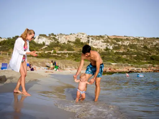 Familia disfrutando de un día en la playa en Menorca con niños. El padre ayuda a su bebé a dar sus primeros pasos en el agua, mientras la madre observa sonriente.