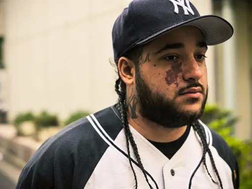 Mężczyzna w czapce z daszkiem i w bluzie z kapturem, z tatuażami na twarzy i szyi, przypominający a$ap yams.