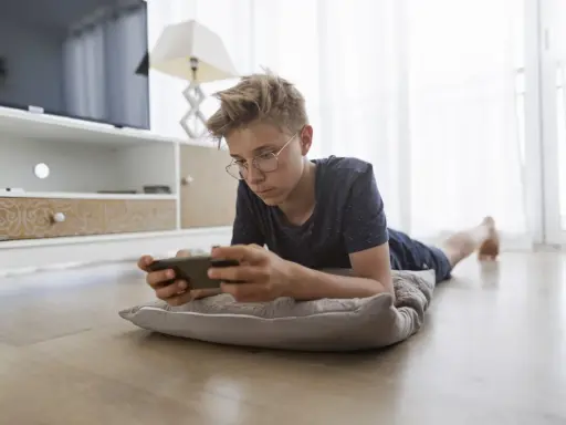Un adolescent, absorbé par son téléphone, illustre la dépendance aux écrans. Il est allongé sur un coussin, le regard fixé sur l'appareil.