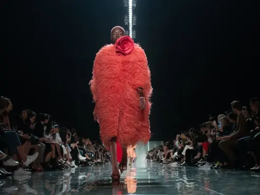 Modella con cappotto vaporoso color corallo e fiore rosso, sfila su una passerella lucida. Il pubblico ammira gli abbinamenti audaci.