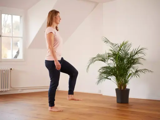 Frau übt auf einem Bein stehen im hellen Raum, neben einer grünen Palme.