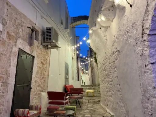 Un vicolo suggestivo nel Val d'Itria, illuminato da lucine, con sedute colorate e muri in pietra bianca.