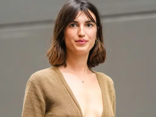 Mujer con cabello castaño ondulado, en transición de pelo corto a largo, luce un suéter marrón y un collar con cruz.