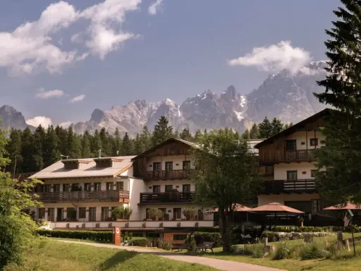 Un hotel alpino immerso nel verde, con imponenti vette innevate sullo sfondo, un perfetto scenario da montagna d'estate.