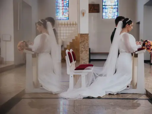 Para młoda klęczy przy ołtarzu. W tle witraże i ławki. Czyżby to były przesądy ślubne dla pana młodego?