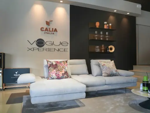 Divano componibile grigio chiaro con cuscini decorativi, logo Calia Italia e "Vogue Xperience" sulla parete.