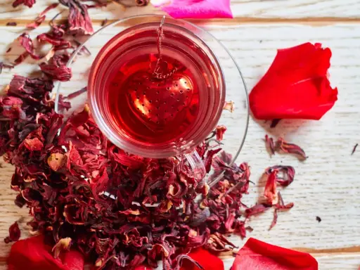 Czerwona, aromatyczna herbata z hibiskusem w szklance z zaparzaczem w kształcie serca, otoczona suszonymi kwiatami i płatkami róż.