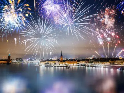 Fyrverkerier lyser upp himlen över Stockholm vid nyår. Vatten reflekterar ljusen från staden och fyrverkerierna.