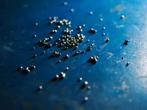 Piccole perle di argento riciclato sparse su una superficie blu scuro, con riflessi luminosi.
