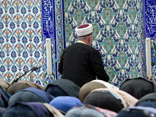 Männer in einer Moschee, die sich im Gebet verneigen. Der Imam trägt einen weißen und roten Hut. Die Fliesenwand ist mit islamischer Kalligrafie und Mustern verziert. Dies ist ein Ort, an dem der größte Sünde im Islam, Shirk, vermieden wird.