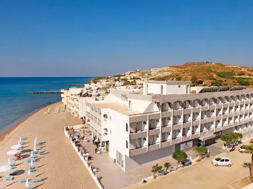 Wypoczynek na plaży w hotelu Island Maya Resort. Biały budynek hotelowy z balkonami, obok piaszczysta plaża z leżakami i parasolami, morze i wzgórza w tle.