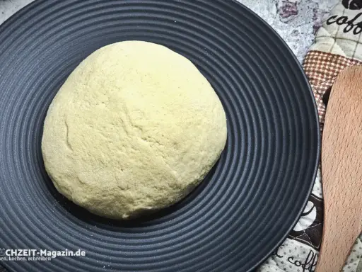 Ein runder, heller pizzateig italienischer Art liegt auf einem schwarzen Teller. Daneben ein Holzspatel und ein Küchentuch.