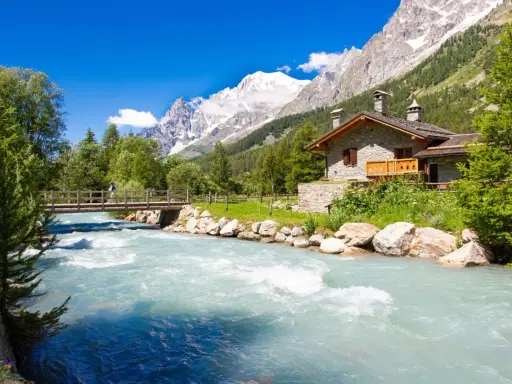 Val Ferret: un'incantevole casa in pietra alpina vicino a un fiume turchese, con un ponte e montagne innevate sullo sfondo.
