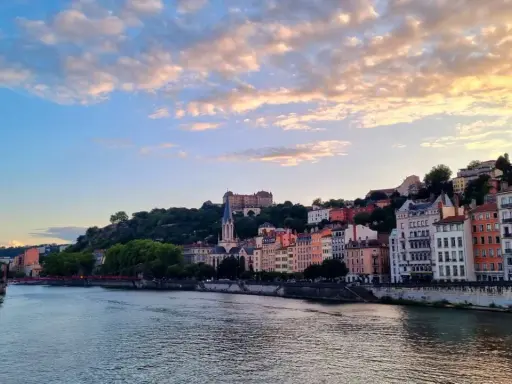 Blick auf die Stadt Lyon, Frankreich, mit dem Fluss Saône im Vordergrund und bunten Gebäuden, die sich den Hügel hinaufziehen. Ein Schloss thront über der Stadt.