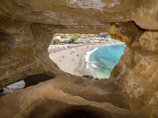 Blick durch Höhle auf Strand von Matala, Kreta. Ein sonniges Hippie-Dorf mit bunten Häusern und blauem Meer.