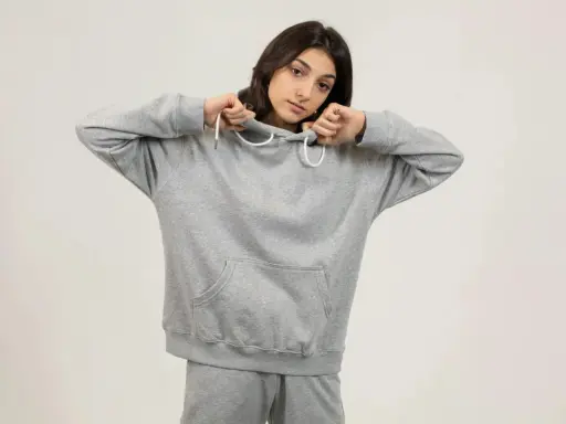 Dziewczyna w szarej bluzie oversize, z kapturem naciągniętym na głowę, pokazuje, jak nosić ten wygodny element garderoby.