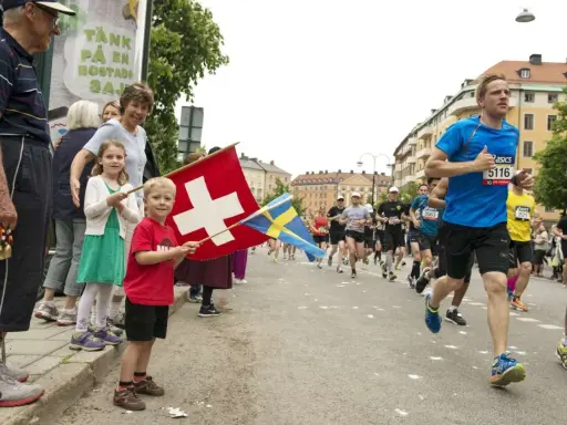 Barn viftar med schweizisk och svensk flagga vid ett lopp. Löpare på marathon distans passerar.