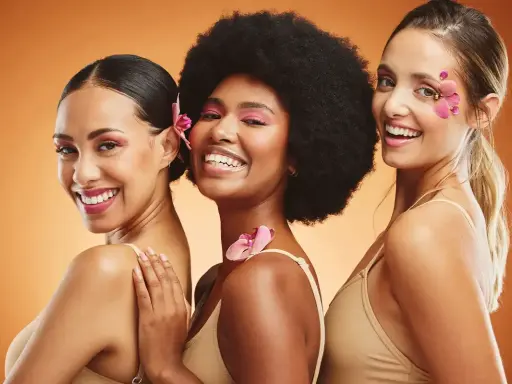 Trois femmes souriantes, un vrai "glow up traduction" avec des fleurs dans les cheveux et un maquillage éclatant.