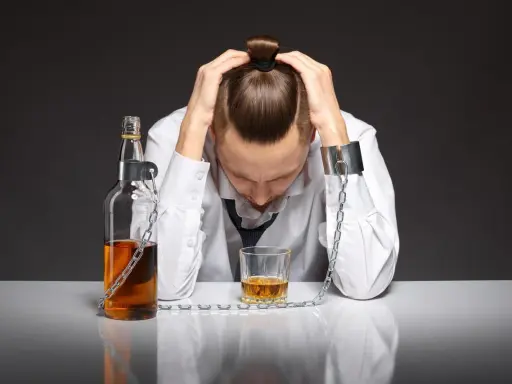 Mężczyzna z rękami na głowie, skuty łańcuchami z butelką i kieliszkiem alkoholu. To symbol walki z alkoholizmem, gdzie pomoc jest kluczowa.