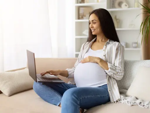 Kobieta w ciąży na kanapie szuka w internecie najdokładniejszy termin porodu, uśmiecha się.
