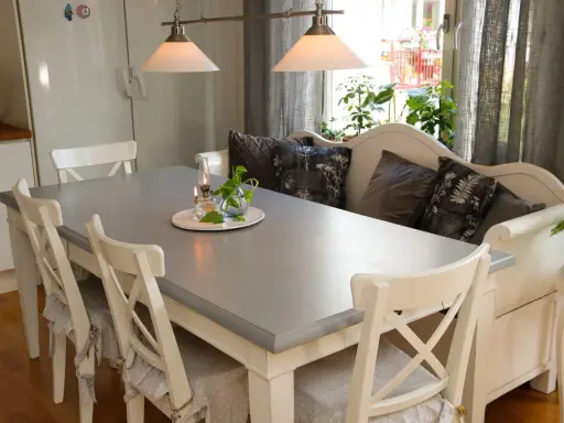 Ett grått bord med vita stolar och en vit bänk med kuddar. Planerar att måla ekbord svart för en modern look.
