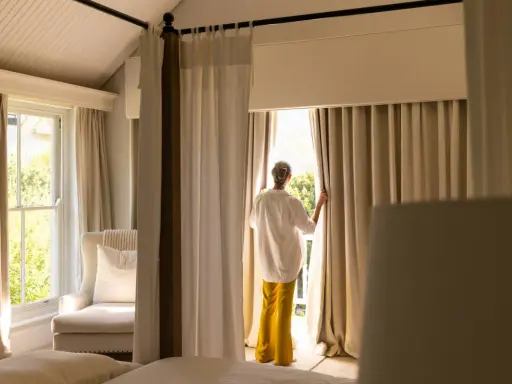 Una mujer abre cortinas para dormitorios, dejando entrar la luz. Se ven camas y un sillón junto a la ventana.