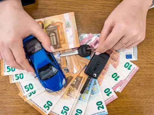 Manos sostienen un coche azul de juguete, llaves y billetes de euro. El coche no arranca, quizás por falta de dinero.