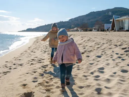 Dwa uśmiechnięte dzieci biegają po piaszczystej plaży. Słońce odbija się od wody, a w tle widać wzgórza i budynek.
