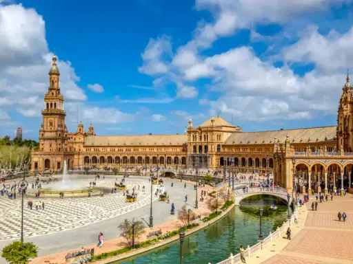 Der beeindruckende Sevilla Palast mit seinen Türmen und Arkaden, umgeben von einem Kanal und belebtem Platz mit Brunnen und Kutschen.